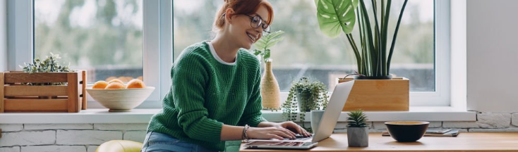 Recarga en línea desde nuestra página fácil y rápido. Chica sentada con suéter verde, jafas y sonriendo usando su laptop para ingresar a Movistar en la sala de su casa.